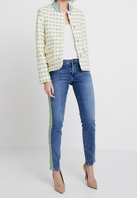 Rautenförmige Tweedjacke in Hellgelb und Blau mit Stehkragen, kombiniert mit blauen Skinny Jeans mit grünen Seitenstreifen.