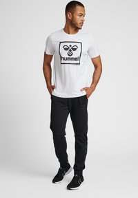 Camiseta blanca con logo negro de "hummel", combinada con pantalones de chándal negros ajustados con detalles laterales y zapatillas deportivas negras.