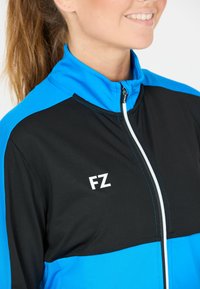 Svart och blå sportjacka med ståkrage, vit dragkedja och FZ-logotyp i vitt på bröstet. Texturerat tyg med kontrasterande sömmar.