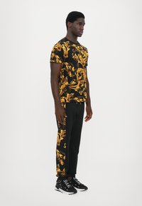 Svart og gullmønstrete t-skjorte med virvlende design; kombinert med svarte bukser med gulldetaljer. Svarte sneakers fullfører stilen.