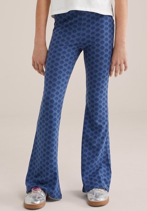 Pantalon évasé bleu à motif floral sombre porté avec des baskets argentées et un haut blanc, visible de la taille aux pieds.