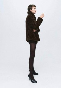 Cappotto in pelliccia sintetica marrone con colletto alto, abbinato a pantaloni corti neri e collant neri opachi. La modella indossa tacchi alti neri, in posizione profilo su uno sfondo grigio.