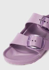Violeta gumijas sandales ar divām pārklājošām siksnām, kurām ir caurspīdīgi plastmasas sprādzes, strukturēts kāju balsts un bieza, veidota zole.