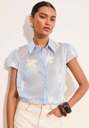Femme aux cheveux foncés en chignon bas, portant un chemisier transparent bleu clair à manches courtes avec broderie florale blanche, pantalon blanc et bracelets épais.