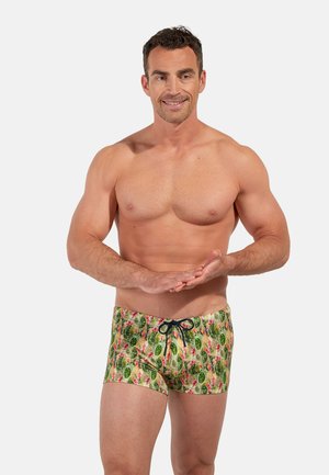 Homme musclé souriant, debout avec les mains jointes devant, portant un short de bain vert avec un motif de cactus et de fleurs.