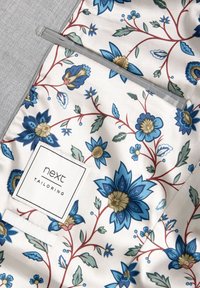 Fodera della tasca floreale con fiori blu e oro su uno sfondo crema. Include un'etichetta bianca con il logo "next tailoring".