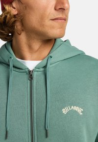Felpa con zip in tessuto verde tenue con coulisse regolabili e logo ricamato "BILLABONG" sul petto, caratterizzata da una texture liscia.