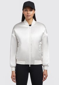 Giacca bomber in raso bianco con chiusura lampo frontale, polsini a costine e due tasche laterali. Il modello indossa un berretto nero e leggins neri.