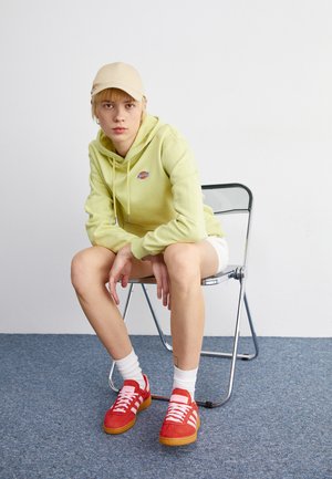 Lys grønn hettegenser med logo, hvite shorts, røde sneakers med hvite striper, sitter på en krom sammenleggbar stol mot en nøytral bakgrunn.