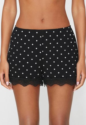 Frau trägt schwarze Shorts mit weißen Punkten und Spitzenbesatz, von der Taille bis zur Mitte des Oberschenkels vor neutralem Hintergrund gezeigt.