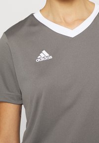 Harmaa urheilupaita, jossa V-aukko ja lyhyet hihat. Valkoinen kaulus ja Adidas-logo vasemmassa rinnassa, valmistettu sileästä, kevyestä kankaasta.