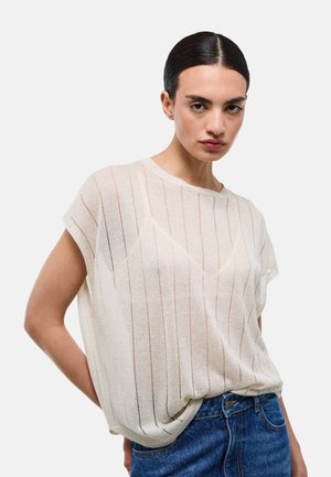 Femme aux cheveux foncés portant un top blanc transparent à manches courtes et un jean bleu, debout avec une main derrière le dos sur un fond uni.