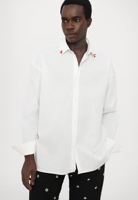 Cămașă albă cu guler, cu o croială lejeră, având detalii de accent roșu pe guler. Asortată cu pantaloni negri decorați cu cuie metalice.
