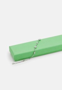 Pulseira prateada com pedras redondas e transparentes, colocada sobre uma caixa verde texturizada. A pulseira tem um design delicado com colocação alternada das pedras.