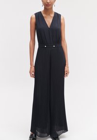 Zwarte geplooide jumpsuit met een V-hals, mouwloos ontwerp en een tailleband met twee knoopaccenten. Gladde textuur met vloeiende stof.