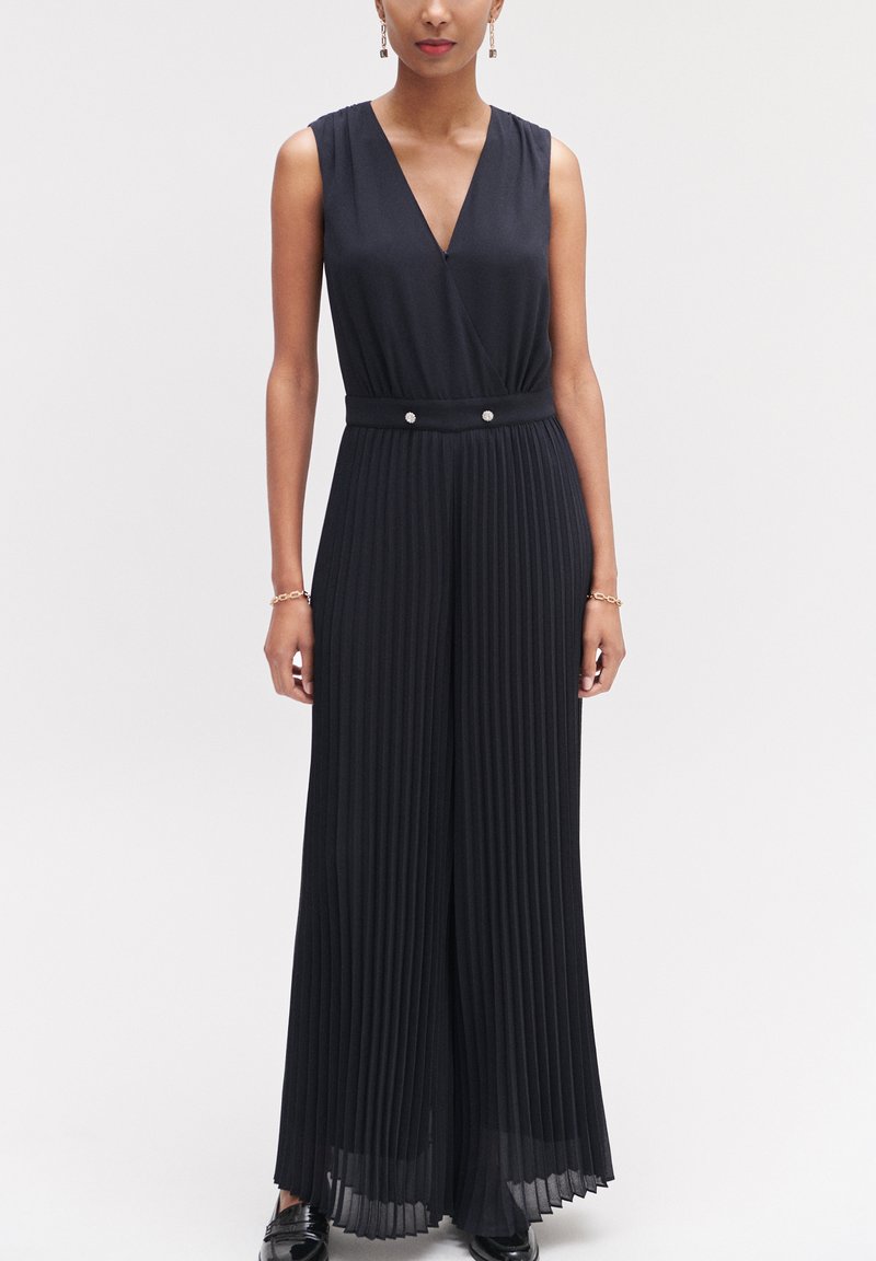 Zwarte geplooide jumpsuit met een V-hals, mouwloos ontwerp en een tailleband met twee knoopaccenten. Gladde textuur met vloeiende stof.
