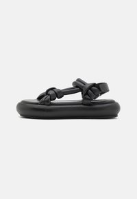 Black sandals with a flat, cushioned sole. Features woven straps and a hook-and-loop fastening for adjustable fit. Smooth leather texture.
