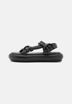 Black sandals with a flat, cushioned sole. Features woven straps and a hook-and-loop fastening for adjustable fit. Smooth leather texture.