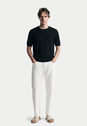 Junger Mann steht mit den Händen in den Taschen, trägt schwarzes kurzärmeliges Hemd, weiße Hose und beige Slide-Sandalen vor weißem Hintergrund.