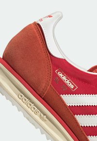 Lähikuva punaisista Adidas SL 72 -tennareista, joissa on valkoiset raidat, beiget pohjat, mokkanahka kantapää ja Adidas-logo kantapäässä ja sivussa.