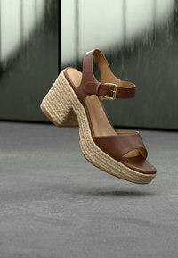 Sandales compensées en cuir marron avec bride à la cheville et semelle en corde tressée flottant au-dessus d'une surface grise avec un fond sombre flou.