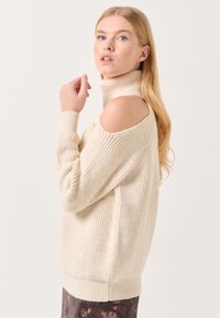 Pull en tricot crème avec un col haut, des découpes aux épaules, une texture côtelée et des manches longues. Présente des fentes sur les côtés pour faciliter les mouvements.