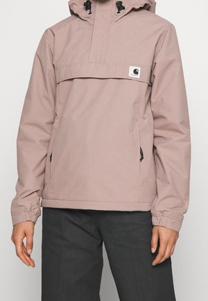Jasnofioletowa kurtka z kapturem, przednią kieszenią z patką, elastycznymi mankietami i logo Carhartt na piersi, noszona z czarnymi spodniami.