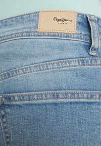 Šviesiai mėlynos džinsinės kelnės su matomu siuvimu, odiniu logotipo etikete "Pepe Jeans London" ir lygiu tekstūros paviršiumi.