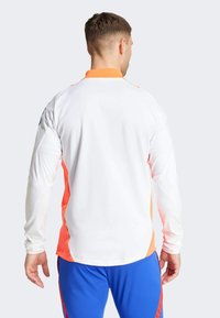 Maglietta atletica a maniche lunghe con corpo bianco, dettagli arancioni e colletto a contrasto; texture liscia; vestibilità rilassata; nessun marchio visibile.