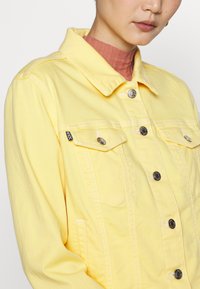 Giacca in denim giallo con colletto classico, tasche frontali e bottoni a pressione argentati. Presenta cuciture in vista e un'etichetta sulla tasca sinistra.
