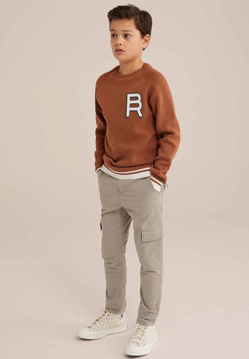 Maglione di colore marrone a maglia con una toppa bianca a forma di "R", abbinato a pantaloni cargo di colore beige chiaro. Scarpe da ginnastica alte bianche completano l'outfit.