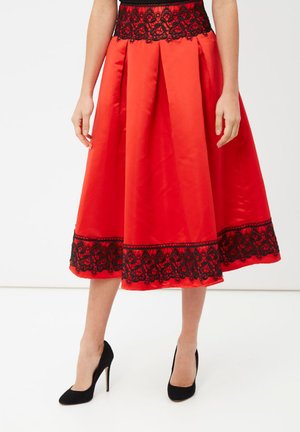 Falda midi roja con ribete de encaje negro en la cintura y el dobladillo, diseño plisado, usada con zapatos de tacón alto negros, de pie sobre fondo blanco.