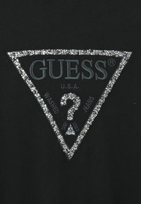 Tissu noir avec un logo Guess triangulaire entouré de perles blanches et argentées, présentant un point d'interrogation en perles et le texte "Washed Jeans".