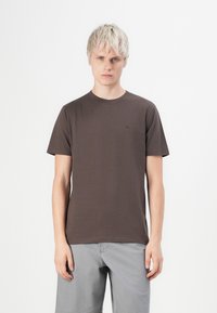 Junger Mann mit kurzem blonden Haar, der ein schlichtes dunkelbraunes T-Shirt und hellgraue Hosen vor einem weißen Hintergrund trägt.