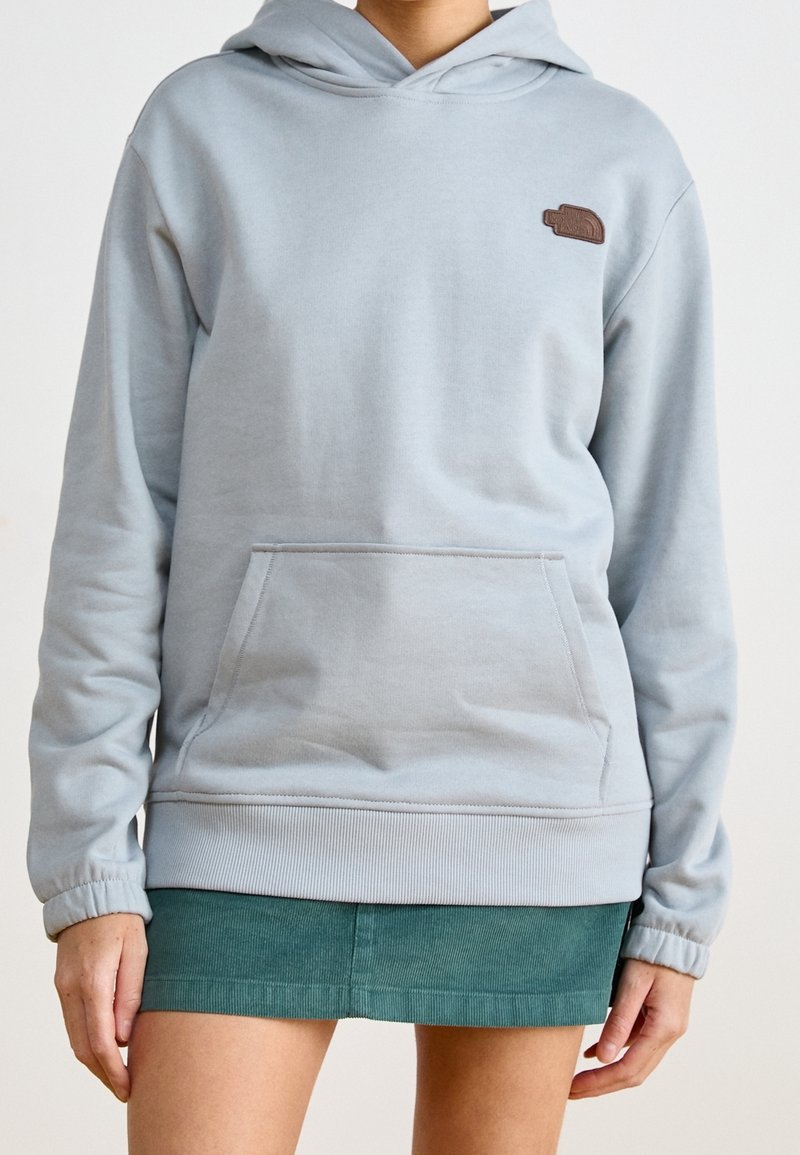Personne porte un sweat à capuche gris clair avec poche avant et petit logo marron, associé à une mini-jupe en velours côtelé verte.