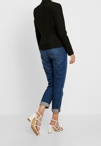 Black tailored blazer, denim cuffed jeans, and white strappy heeled sandals. The jacket has a fitted design, smooth texture, and structured shoulders.