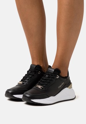 Zapatillas de cuero negro con un cuello acolchado, detalles de hardware dorado y una gruesa suela blanca. Presenta un logo en el lateral y una parte superior texturizada.