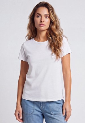 Femme aux longs cheveux ondulés portant un t-shirt blanc uni à manches courtes et un jean bleu clair, debout devant un fond neutre.