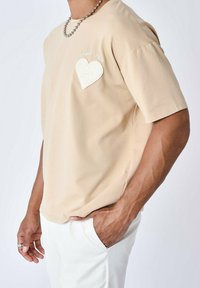 Camiseta de manga corta beige en tejido suave con un bolsillo texturizado en forma de corazón blanco y "Project X" bordado encima del bolsillo.