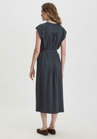 Hame midi harmaja dengan lengan pendek, ikat pinggang, dan detail belakang yang berkumpul. Tekstur halus, desain mengalir, dan potongan sepanjang pergelangan kaki.