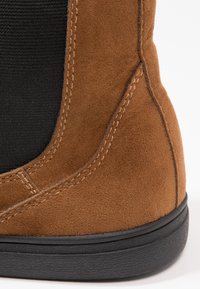 Brązowy zamszowy botek za kostkę z czarną elastyczną wstawką, okrągłym noskiem i gumową podeszwą o fakturowym wykończeniu. Widoczne detale przeszycia.