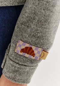 Sivý textilný rukáv s nášivkou znázorňujúcou croissant na levanduľovom károvanom pozadí s žltým slnečným akcentom.