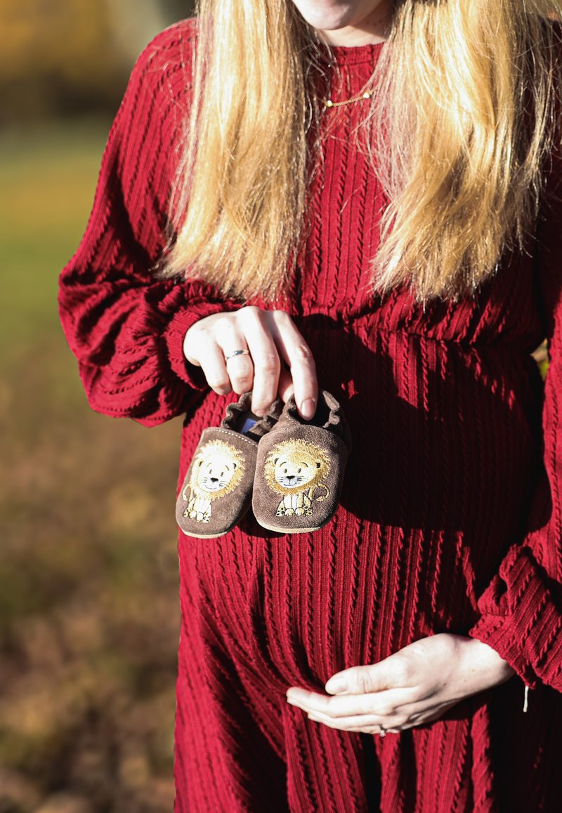 Bruine babyschoentjes met leeuwenpatronen, vastgehouden in één hand door een persoon in een gestructureerde rode jurk die een ronde buik ondersteunt.