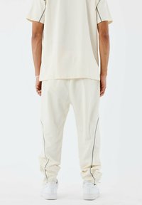 Conjunto deportivo color crema que incluye una camiseta de corte holgado y pantalones tipo tapered con franjas laterales grises, combinado con zapatillas blancas.