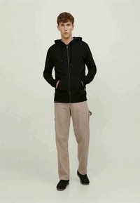 Jovem de pé, vestido com um hoodie preto com fecho, calças bege e sapatos pretos, contra um fundo claro e simples.