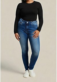 Mujer vestida con una camiseta negra de manga larga, jeans ajustados azules de cintura alta y zapatillas blancas, de pie contra un fondo liso.