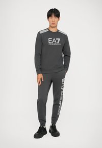 Súprava šedej mikiny a nohavíc s bielym logom "EA7 Emporio Armani", so pruhovanými detailmi na pleciach a pohodlným strihom.