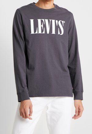 Person iført en mørkegrå langærmet skjorte med hvid "LEVI'S" tekst på brystet og hvide bukser, stående foran en ensfarvet baggrund.