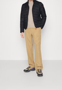 Schwarze Steppjacke, beiger Pullover, braune Hose und mehrfarbige Sneaker. Glatte Textur, vertikales Rautenmuster, orangefarbener Reißverschluss-Anhänger.
