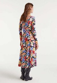 Mujer de espaldas vistiendo un vestido floral de manga larga hasta la mitad de la pantorrilla con flores rojas, moradas, amarillas y blancas, combinado con botas altas negras hasta la rodilla.